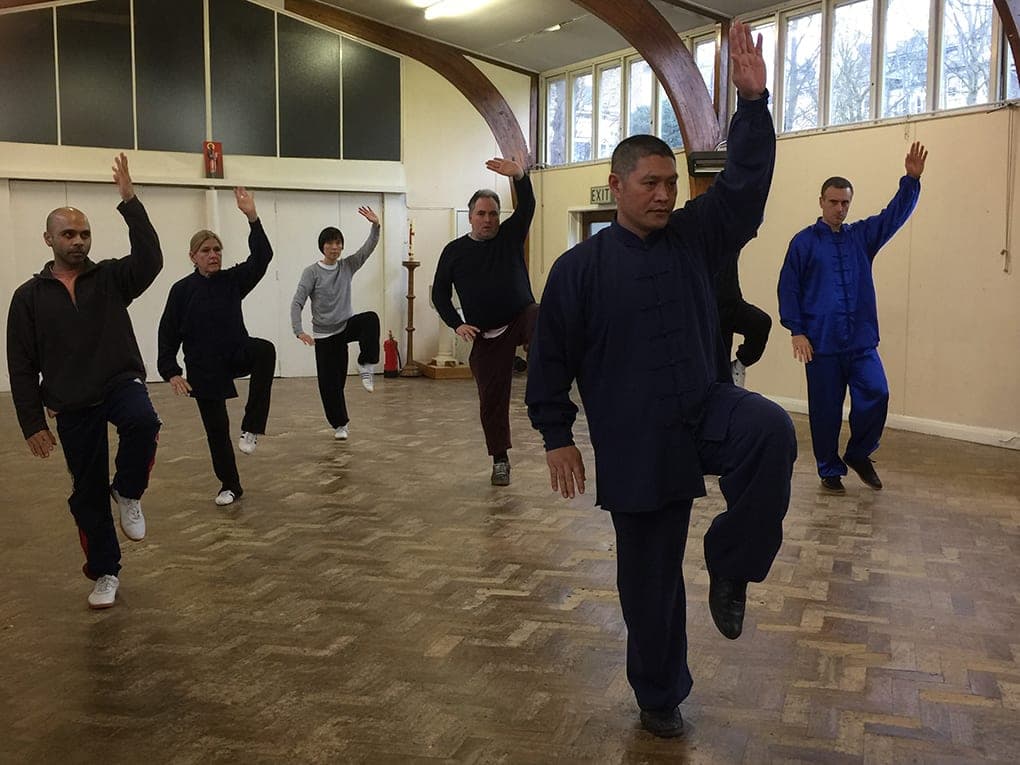 Shifu Liu leads students in a one-legged stance with one arm raised in a parquet-floored hall