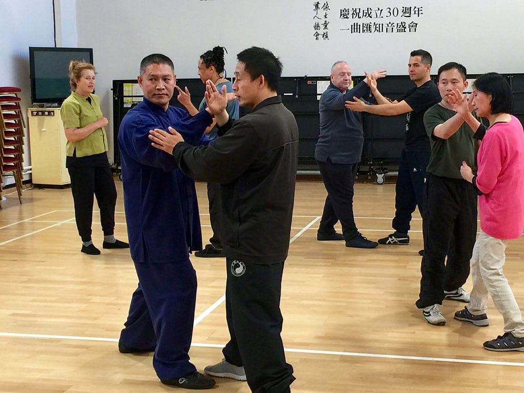 Master Chen Ziqiang and Shifu Liu practise push hands in a sports hall while students pair up behind them