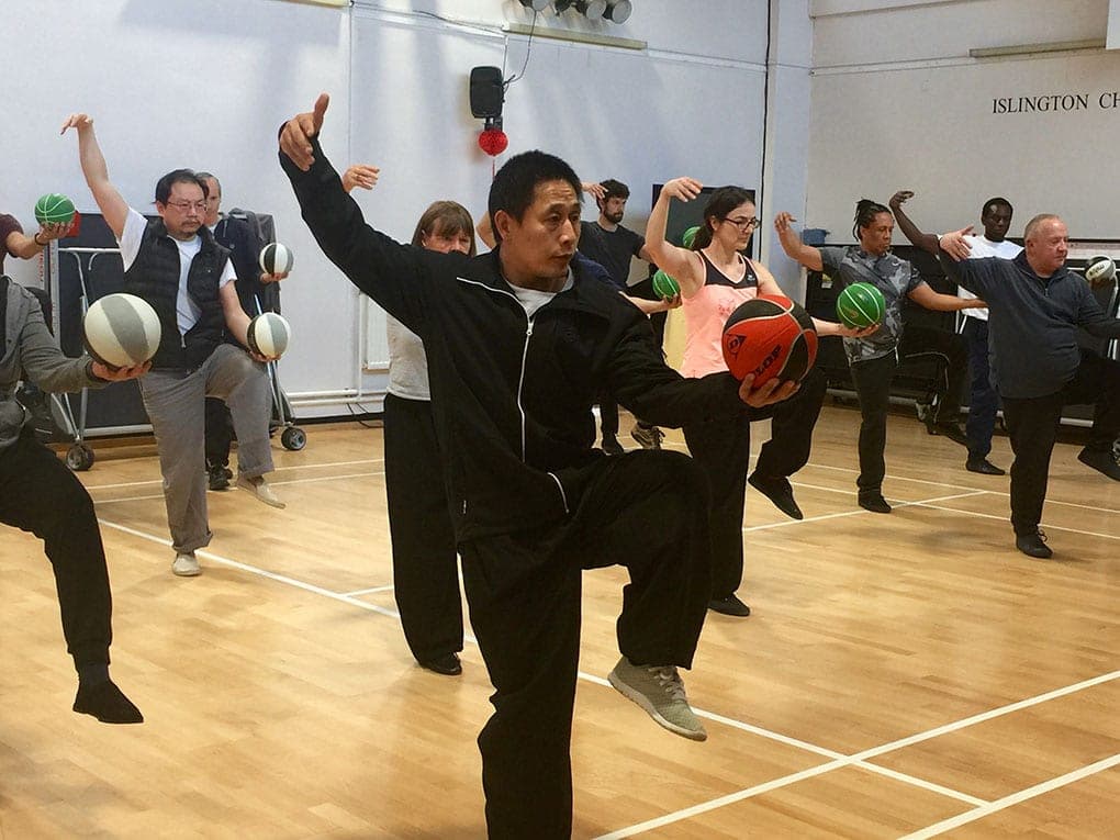 Master Chen Ziqiang, in black, raises a basketball on one knee while students copy the posture with their own balls in a sports hall