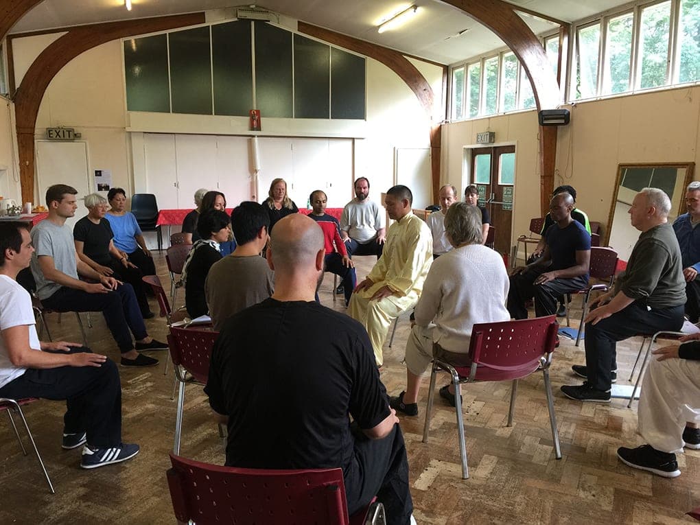 Shifu Liu sits in a circle of students on chairs during a seated Qigong session in a hall
