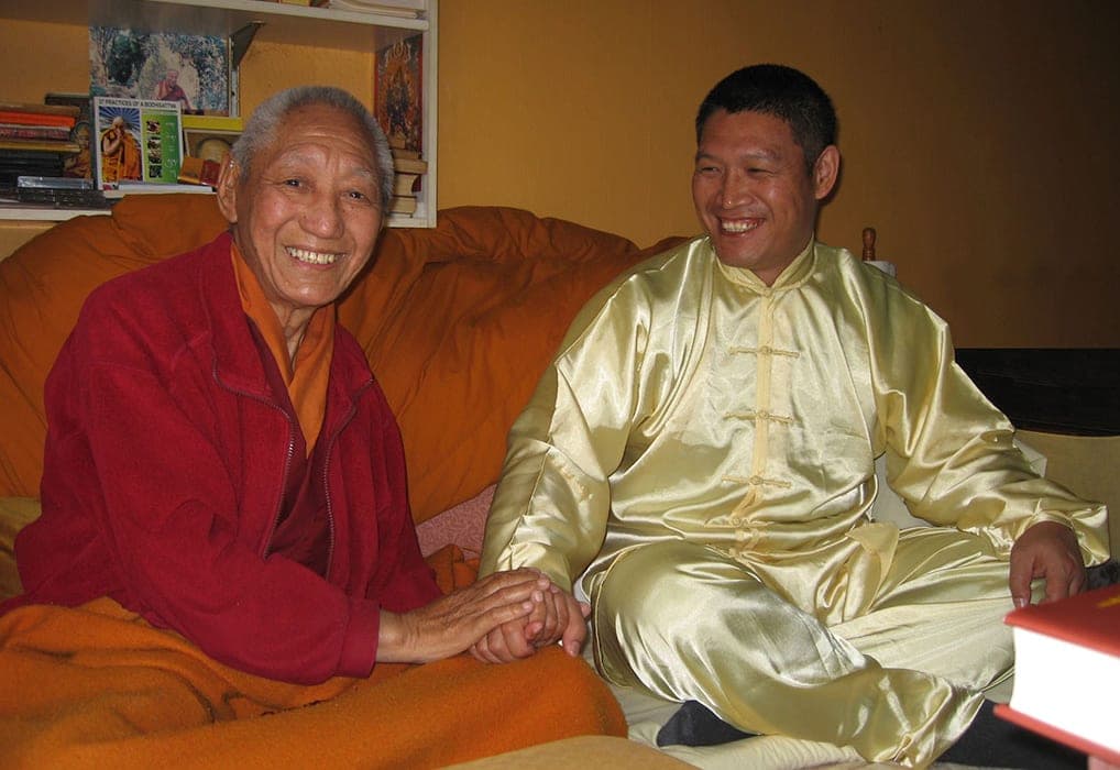 The Venerable Geshe Damcho Yonten, in maroon robes, and Shifu Liu hold hands and smile on a sofa