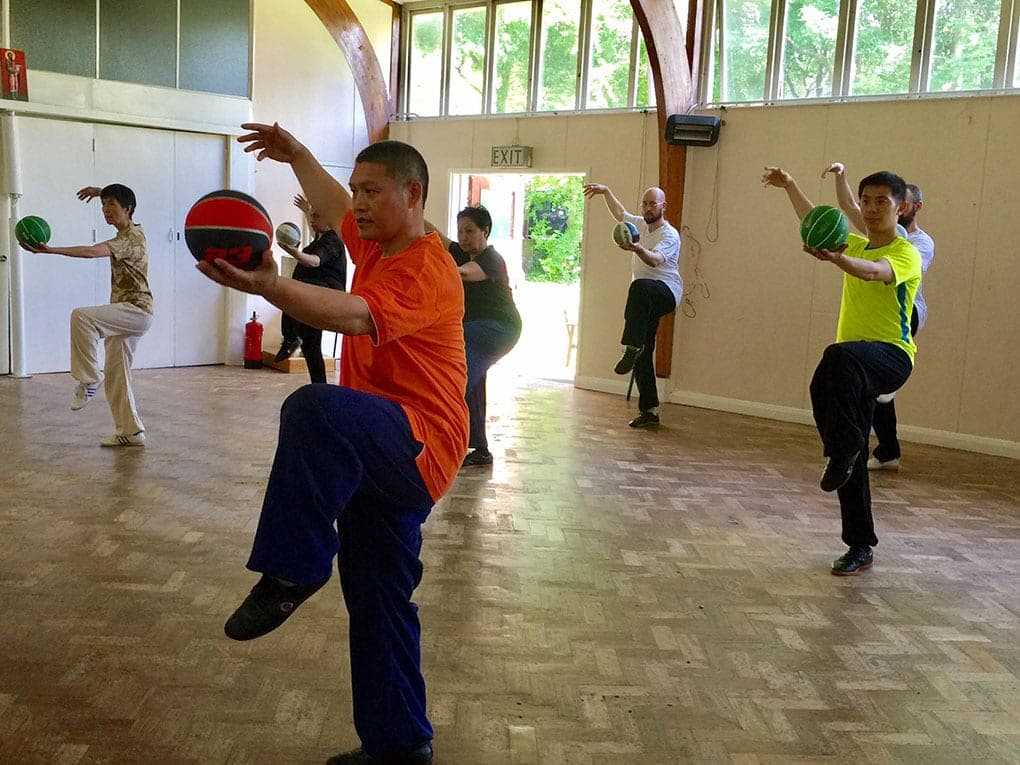 Shifu Liu in an orange T-shirt balances on one leg holding a basketball, with students doing the same behind him