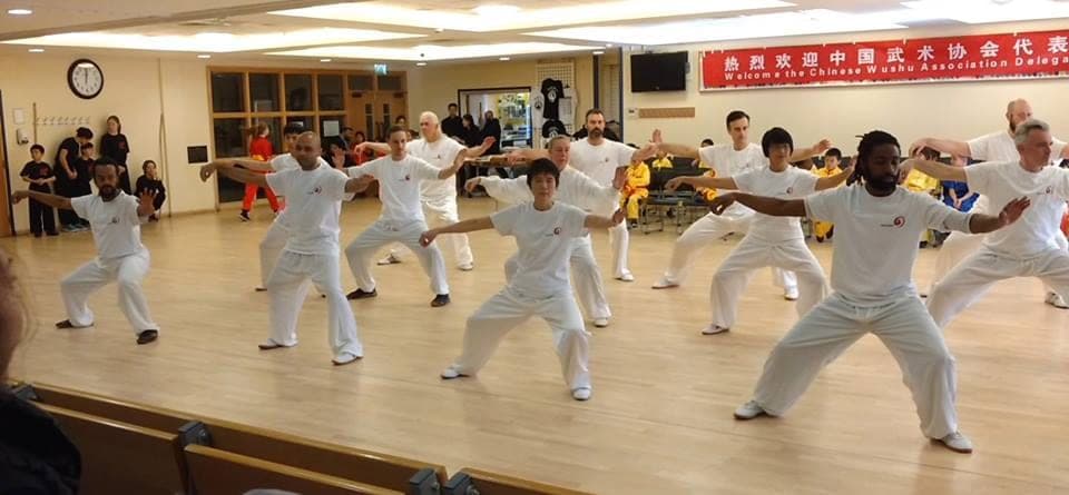 Performers in white T-shirts and trousers practise a Taiji posture in rows in a hall hung with a banner welcoming the Chinese Wushu Association delegation