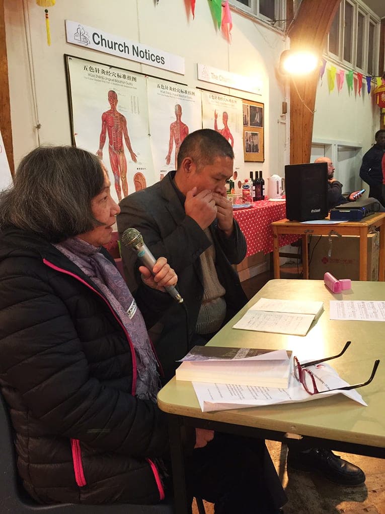 A student sings into a microphone at a table while Shifu Liu plays the harmonica beside her