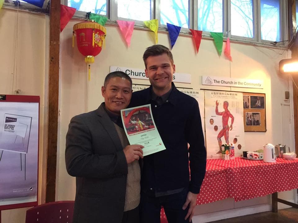 Shifu Liu and a student smile with an assessment award certificate beneath bunting and a red lantern