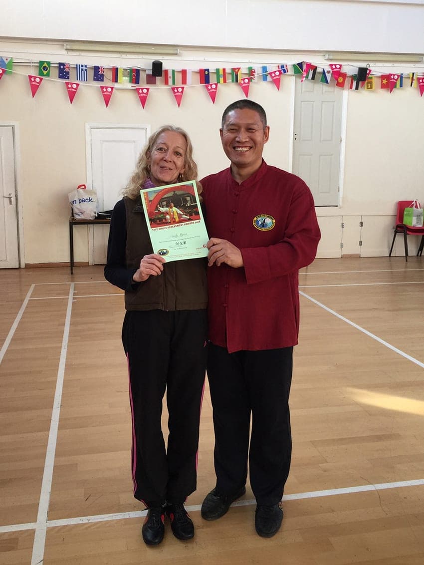Shifu Liu and a student hold her assessment award certificate in a sports hall hung with flags