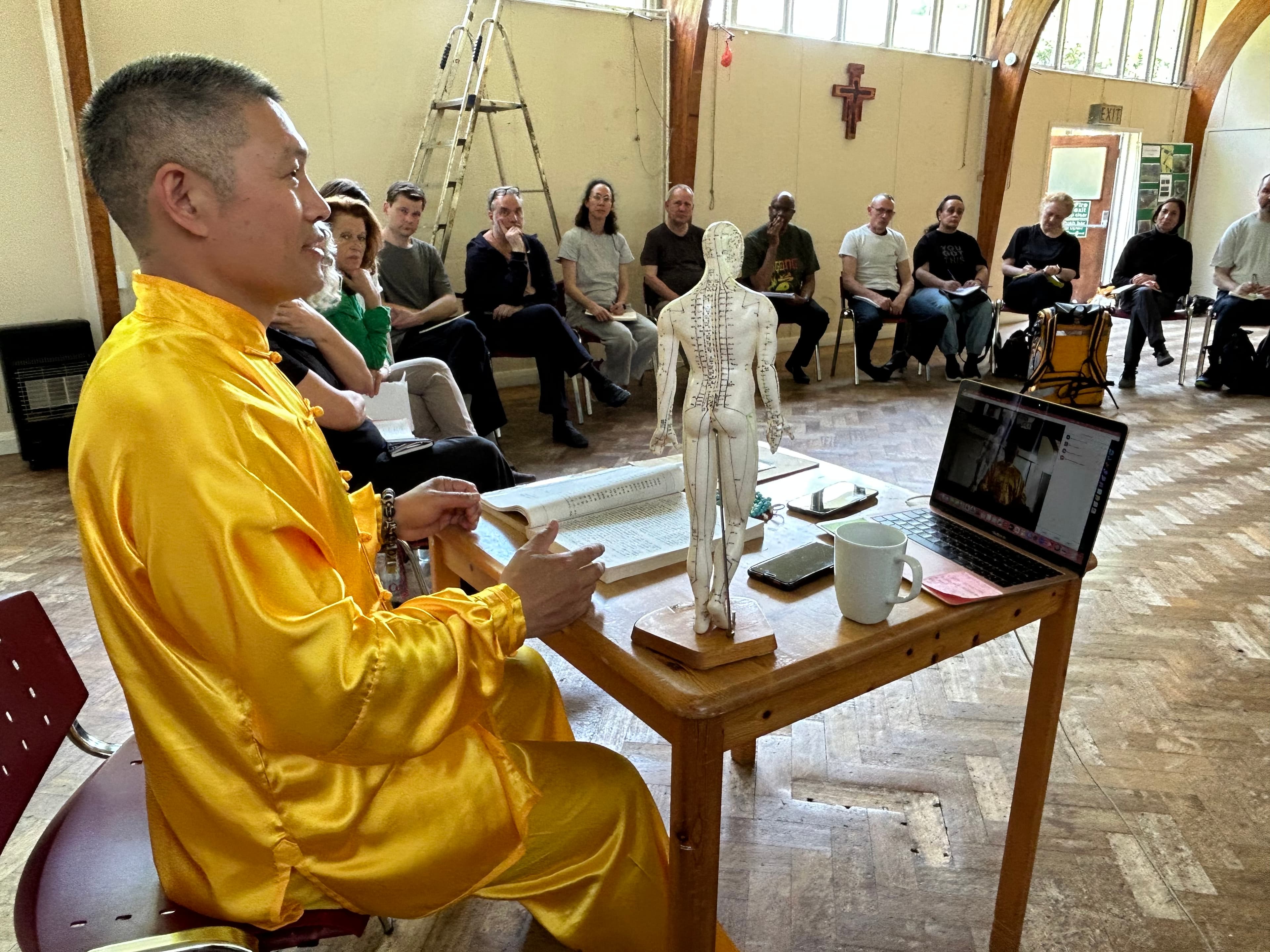 Shifu Liu in gold uniform teaching Traditional Chinese Medicine with an acupuncture model to a seminar audience