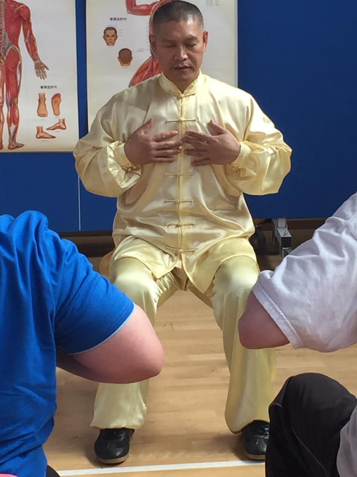 Shifu Liu in gold uniform seated at a desk teaching Traditional Chinese Medicine with anatomy posters behind