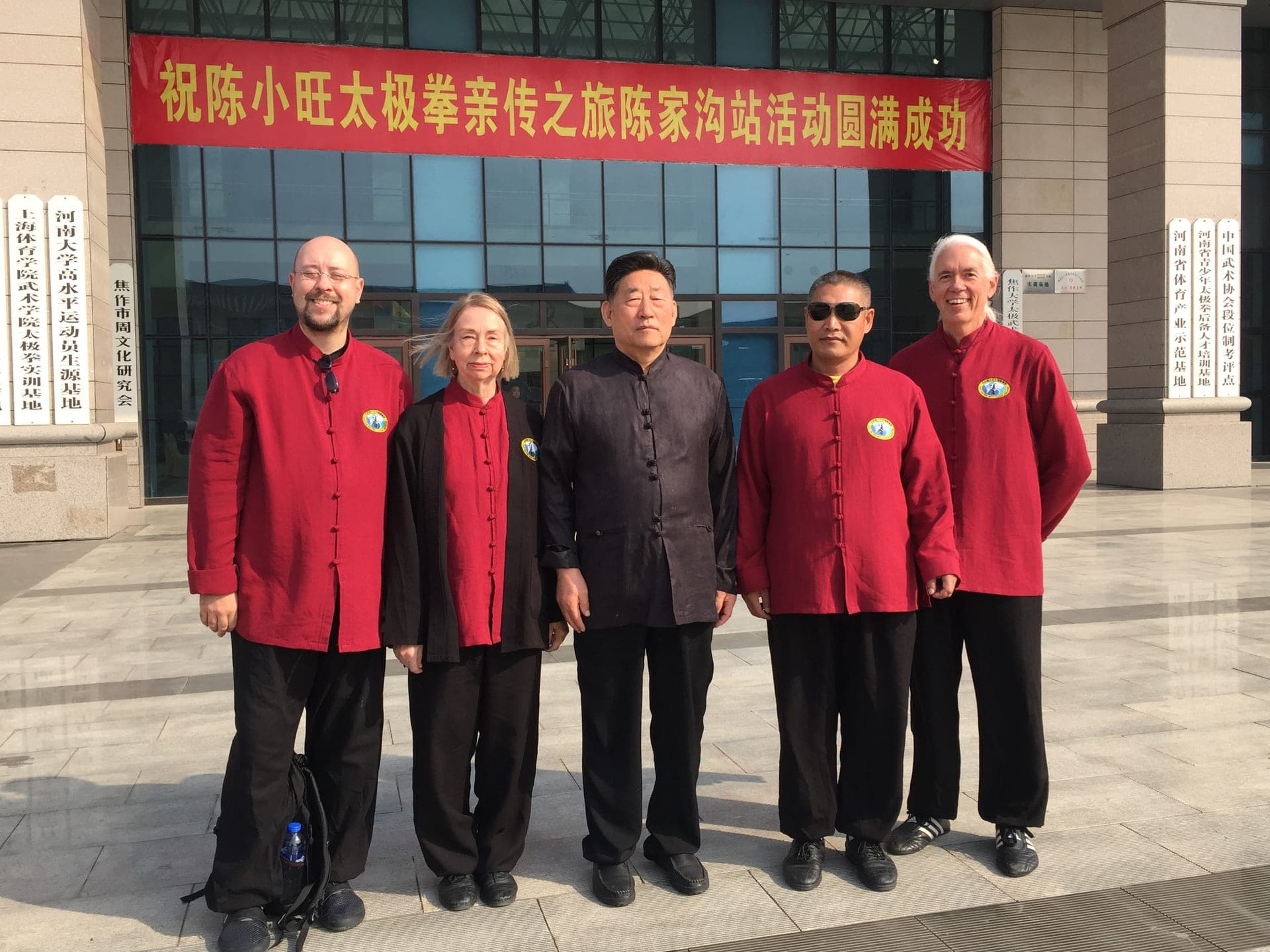 Five people in red and black Chinese-collar jackets stand in front of a building in Chenjiagou, under a long red banner with Chinese writing