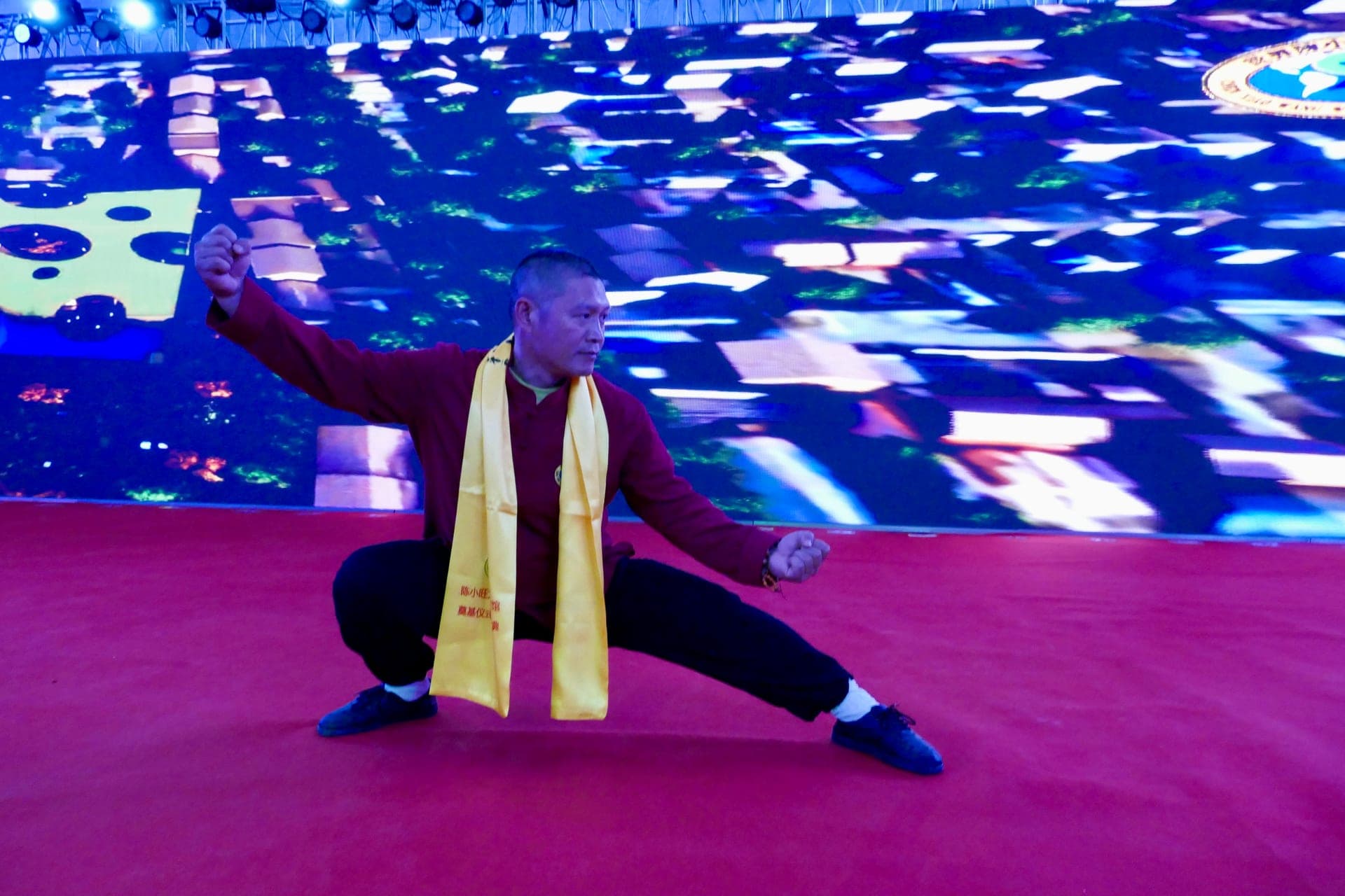 A practitioner wearing a yellow ceremonial sash drops into a deep low stance on a red stage in front of a giant screen