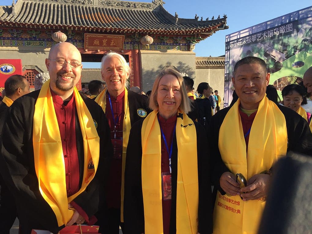 Four people in black jackets and yellow ceremonial sashes smile in front of a temple gate in Chen Village, beside a board showing an aerial view of the planned museum