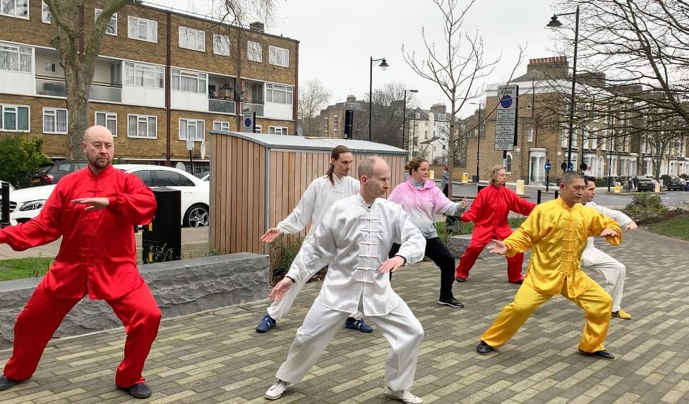 Performers in red, white and yellow silk holding low stances together on a paved square lined with trees and flats