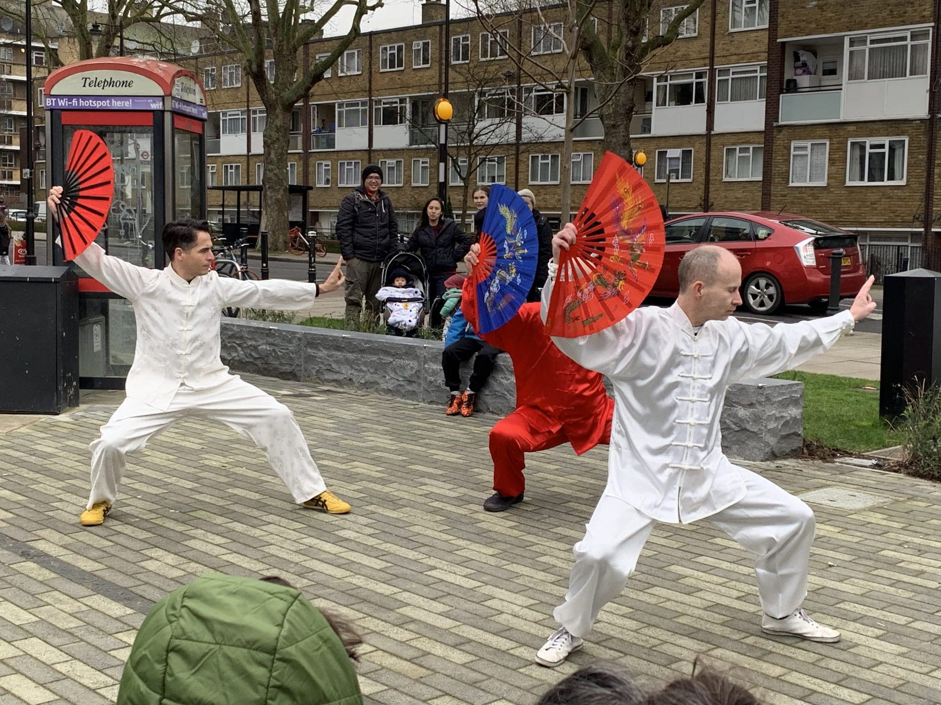 Two performers in white silk opening red fans, with a third in red behind them, as a small crowd watches
