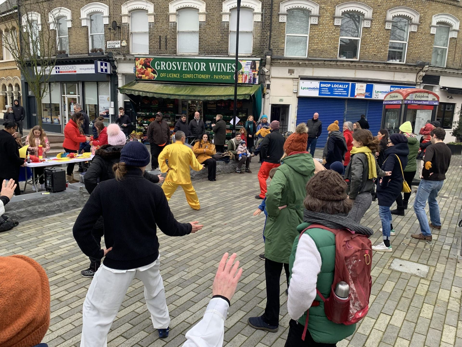 A crowd in coats and hats following the Taiji movements in front of a row of shops