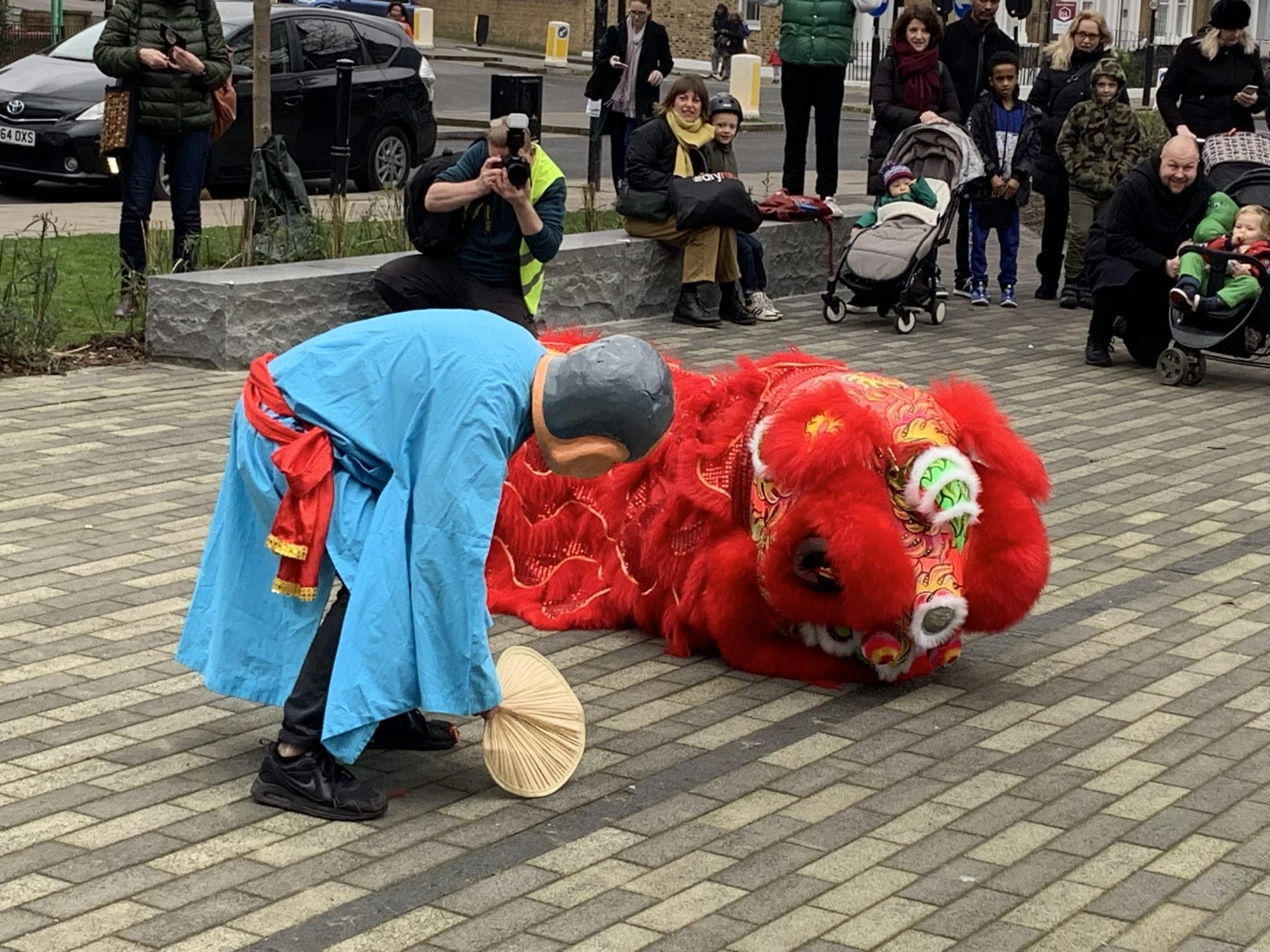 A masked figure in a blue robe bowing with a fan in front of a red lion lying on the paving, with onlookers behind