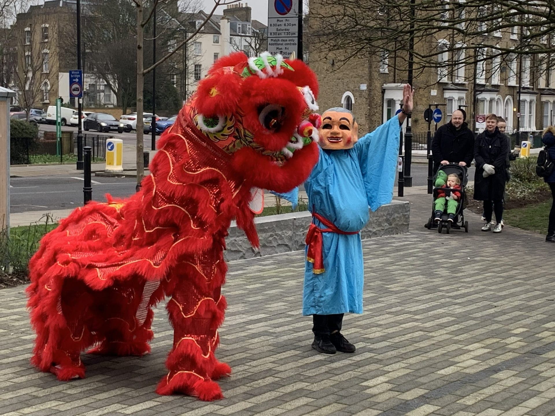 A red Chinese lion rearing up beside a masked figure in a blue robe on the street