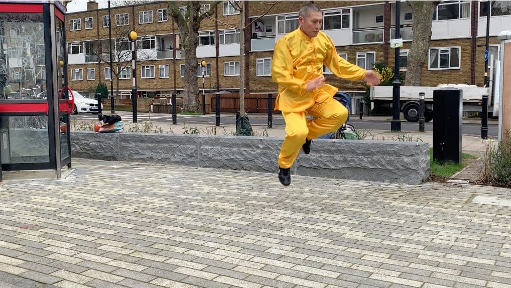 Shifu Liu Quanjun in yellow silk leaping in a jump from Xinjia Yilu on a paved square in Canonbury