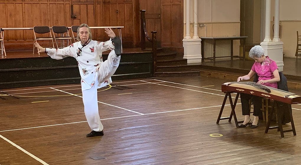 Chris Jones, in a white embroidered jacket, holds a high kick in a wooden-floored hall while a musician plays the guzheng beside her