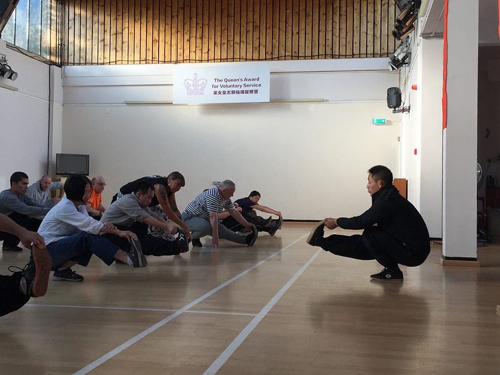 The class sits on the floor of a sports hall stretching forward over their legs as the teacher demonstrates a low squatting stretch