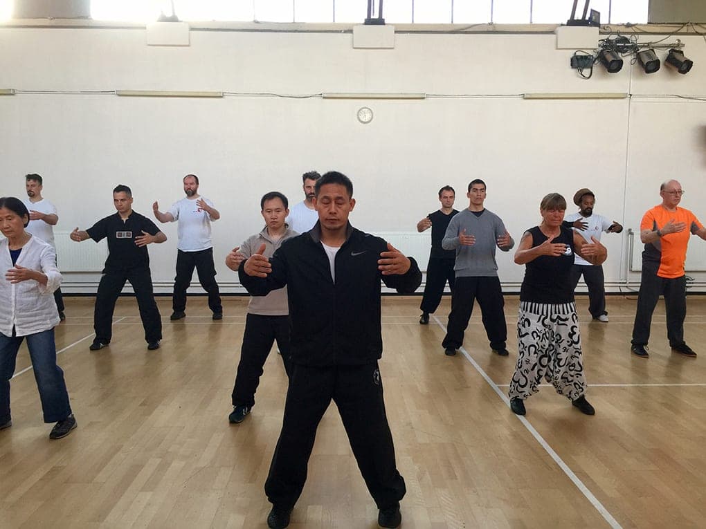 A large class stands in rows holding the Zhan Zhuang standing posture, arms rounded in front of the body, a teacher in black at the front
