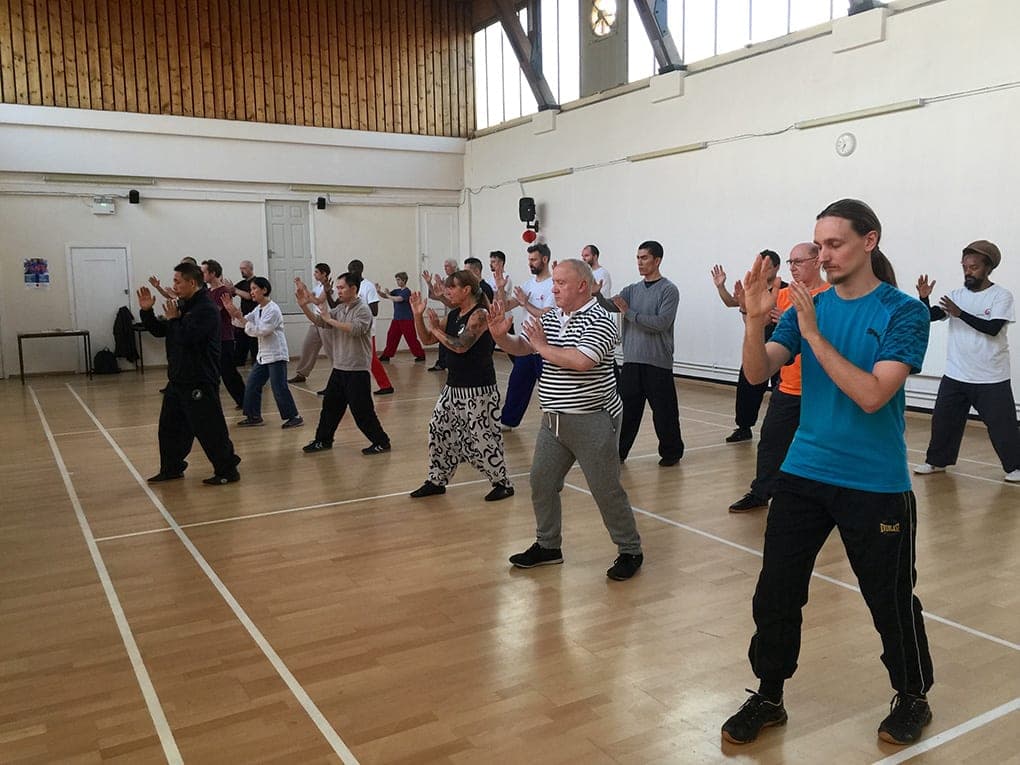Rows of students practising silk reeling across a sports hall, hands raised in front of them