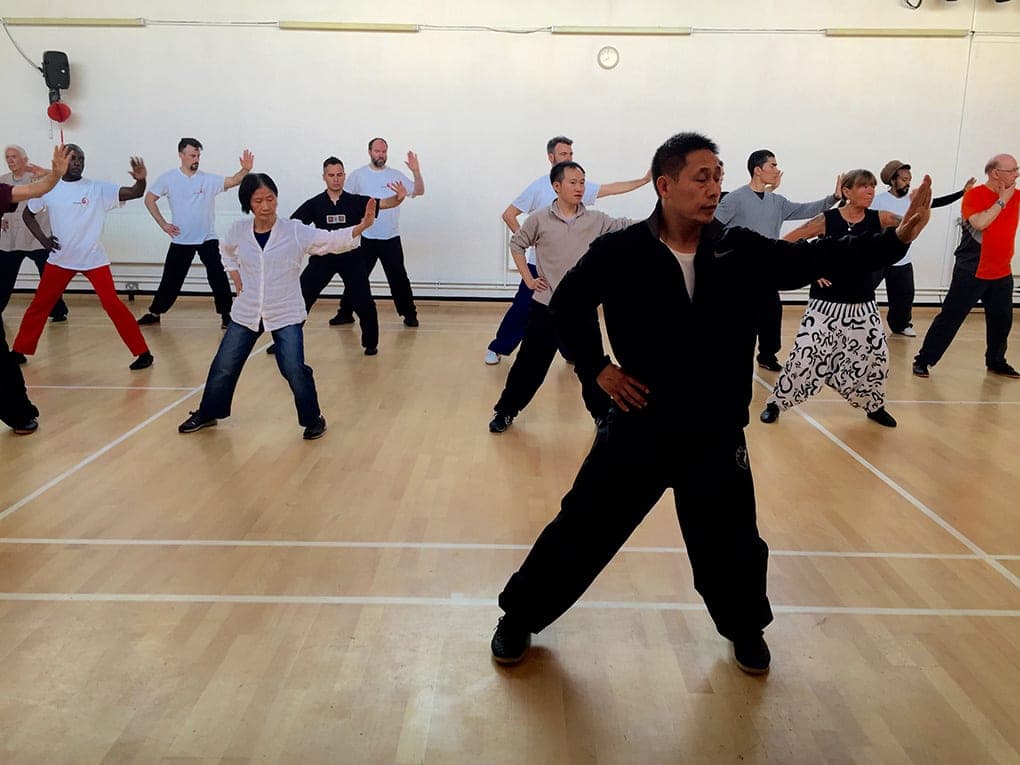 A teacher in a black tracksuit leads a large class through a silk reeling movement in a sports hall, one arm extended