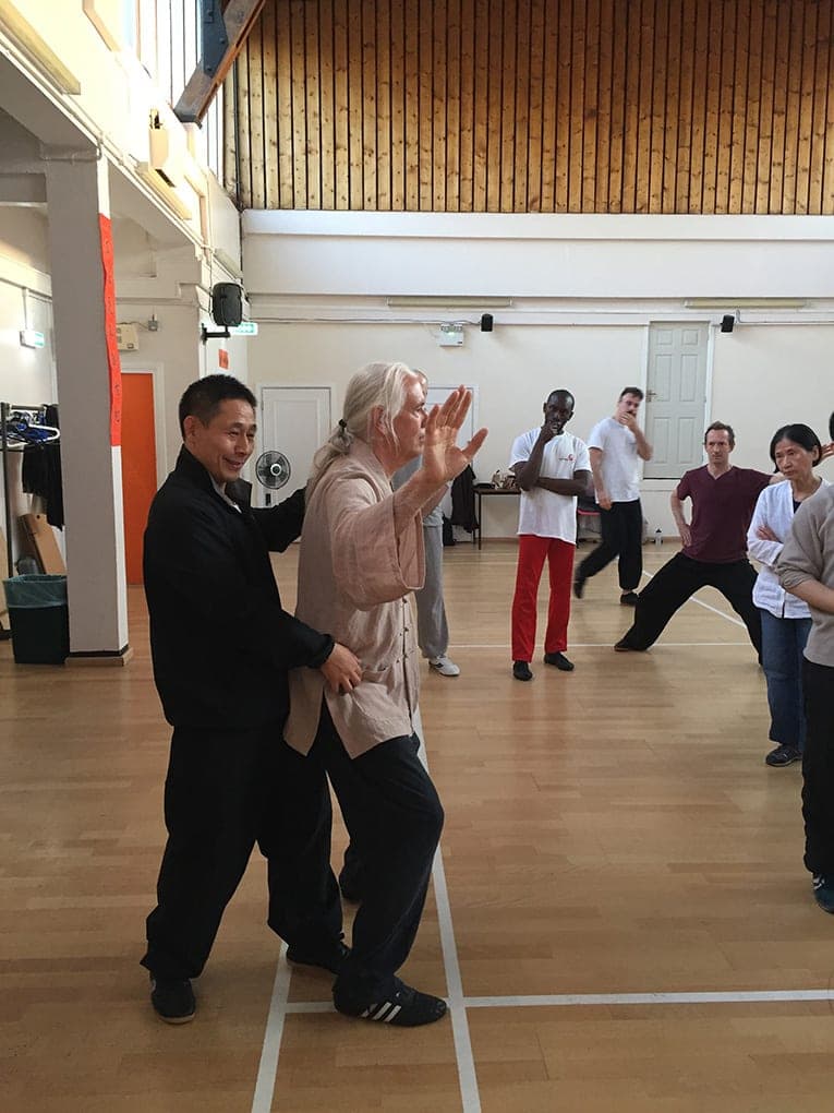 A teacher in black adjusts the posture of a student with long pale hair while the class practises behind them