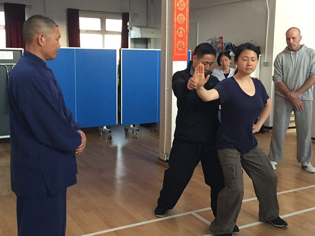 A teacher stands behind a student and adjusts her arm and stance while a man in a blue Chinese-style suit watches