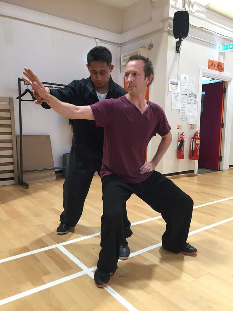 A teacher stands behind a student in a maroon T-shirt and corrects his low stance and outstretched arm