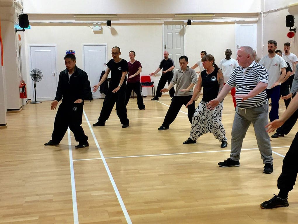 Students in a sports hall step into low stances, arms held out to the side