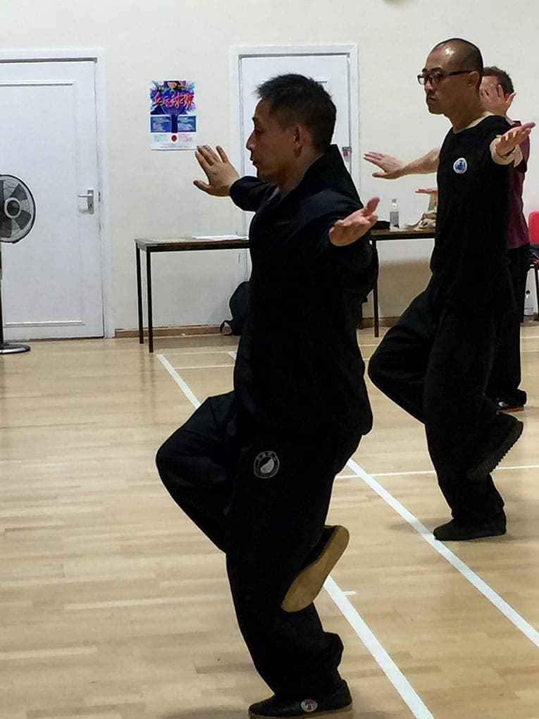 Two men in black practise a raised-knee posture, one close to the camera and one behind