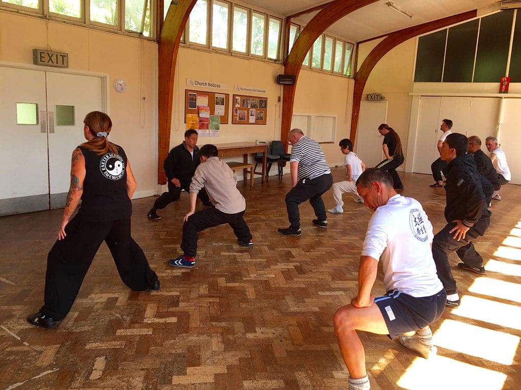 Students in a church hall sink into low warm-up stances on a sunlit wooden floor