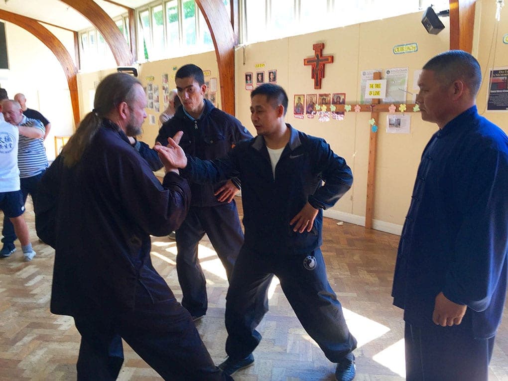 Two men work through a push hands exercise while others look on in front of a wall with a crucifix