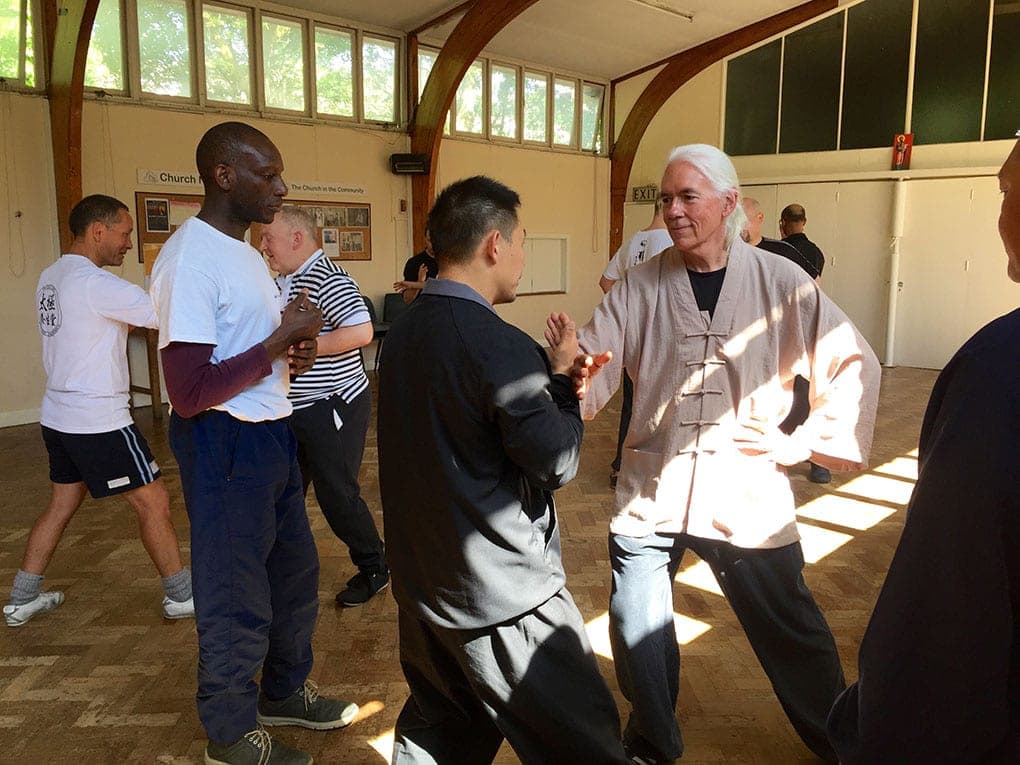 Pairs practising push hands in a sunlit church hall with a herringbone wooden floor