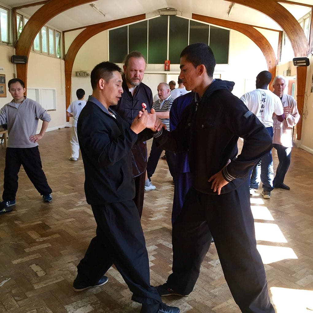 A teacher in a dark tracksuit works with a student in a hooded top while other pairs practise behind
