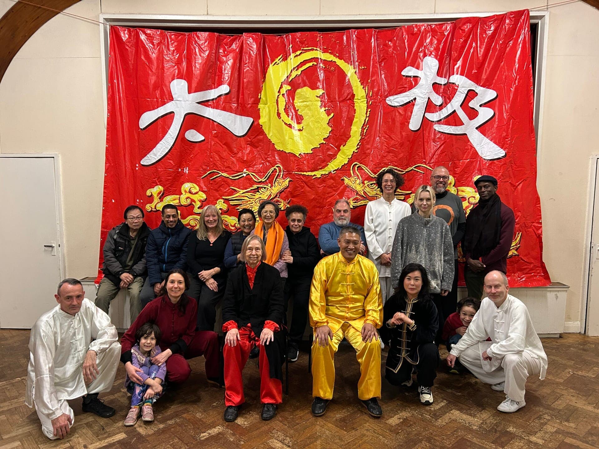 A large group of students, some in white and red practice suits, gathered around a teacher in yellow silk in front of a red banner bearing the characters 太极