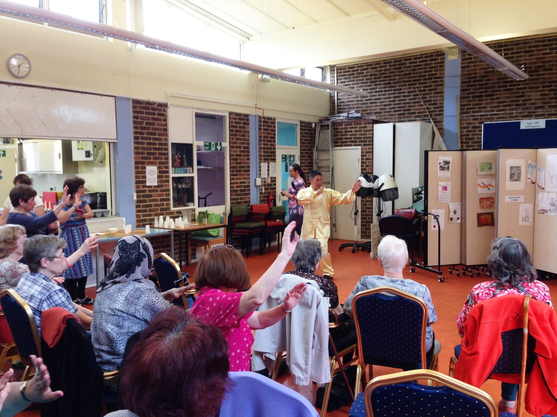 Shifu Liu in yellow silk demonstrates a Taiji movement at the front of a community hall while seated older people follow, raising their arms