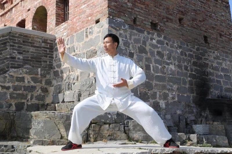 Master Chen Bing in white holding a low Taiji stance in front of an old stone wall