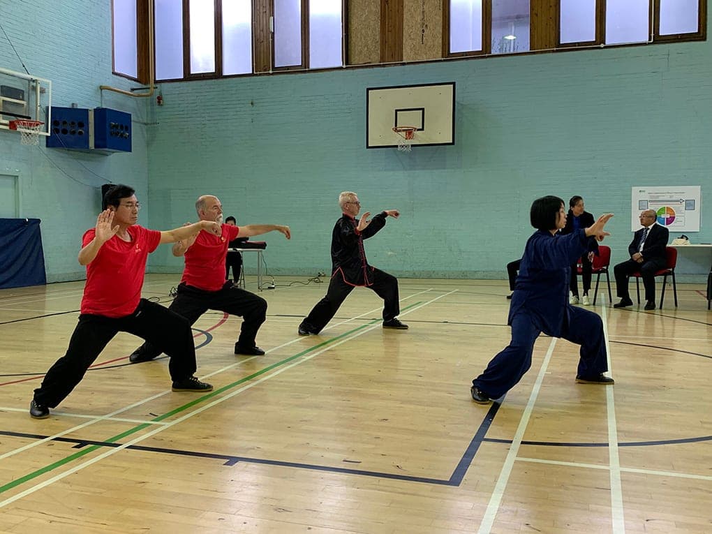 A Taiji group in red, black and blue performing low stances in the Edinburgh sports hall, with Wen Li