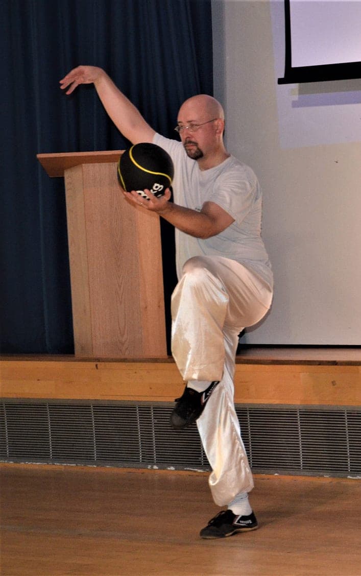 A student in white on one leg holding a Chen Taiji ball at shoulder height