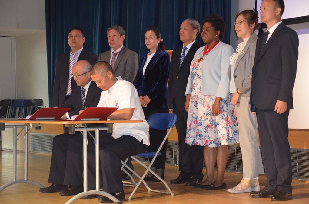 Two men signing documents at a table on stage while Councillor Sabrina Francis and members of the delegation look on