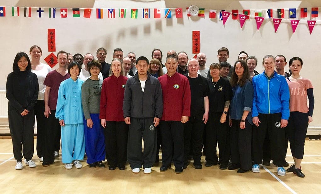 A large group photo of workshop participants standing in rows in a sports hall hung with international flag bunting