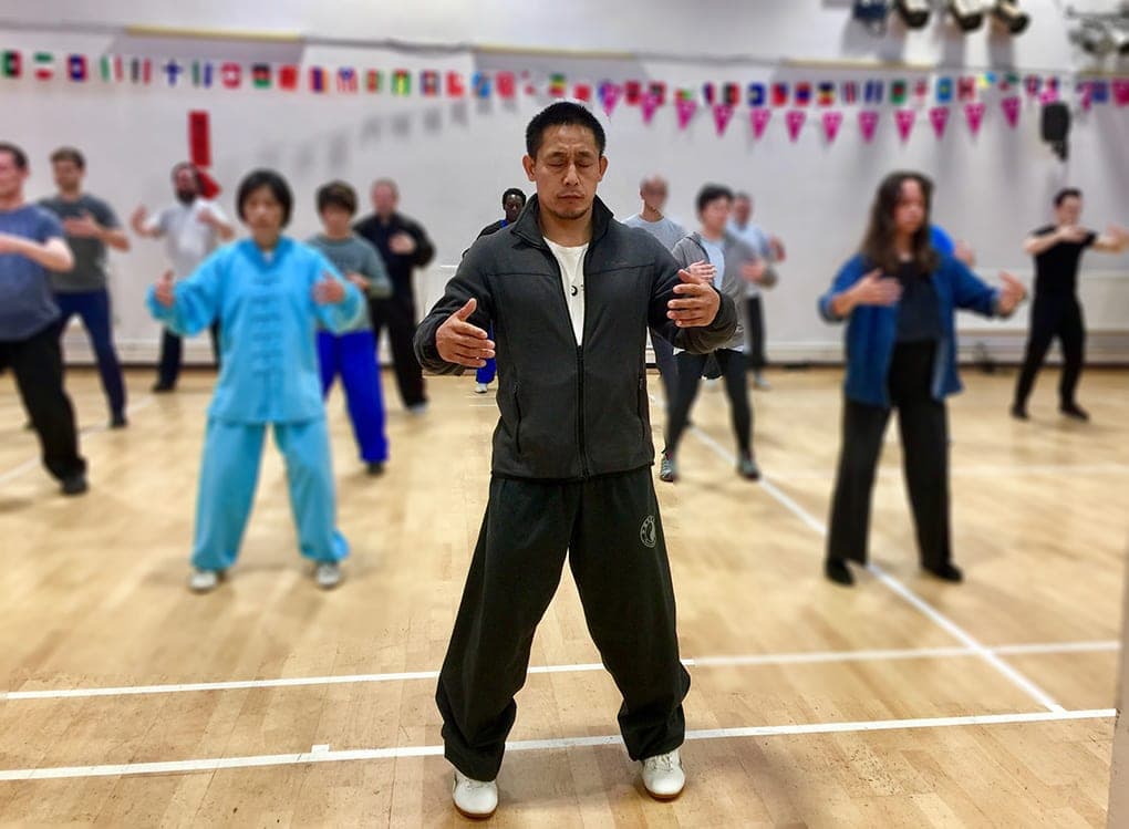 A teacher in a dark tracksuit stands at the front of a sports hall in the Zhan Zhuang posture, arms rounded, with students standing behind him