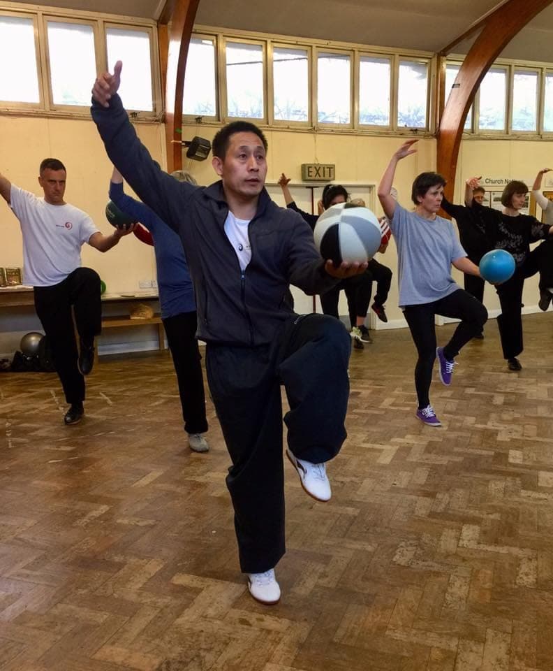 A teacher stands on one leg with one arm raised and a Taiji ball in the other hand, students with balls behind him in a church hall