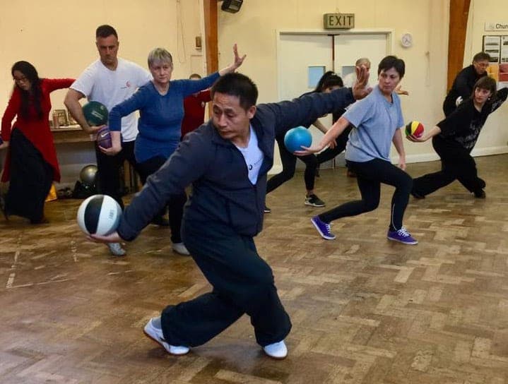 A teacher sweeps low in a long stance holding a ball, with the class following behind him