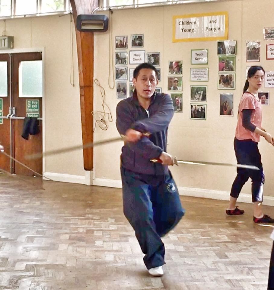 A teacher turns with two swords in motion while a student practises behind him