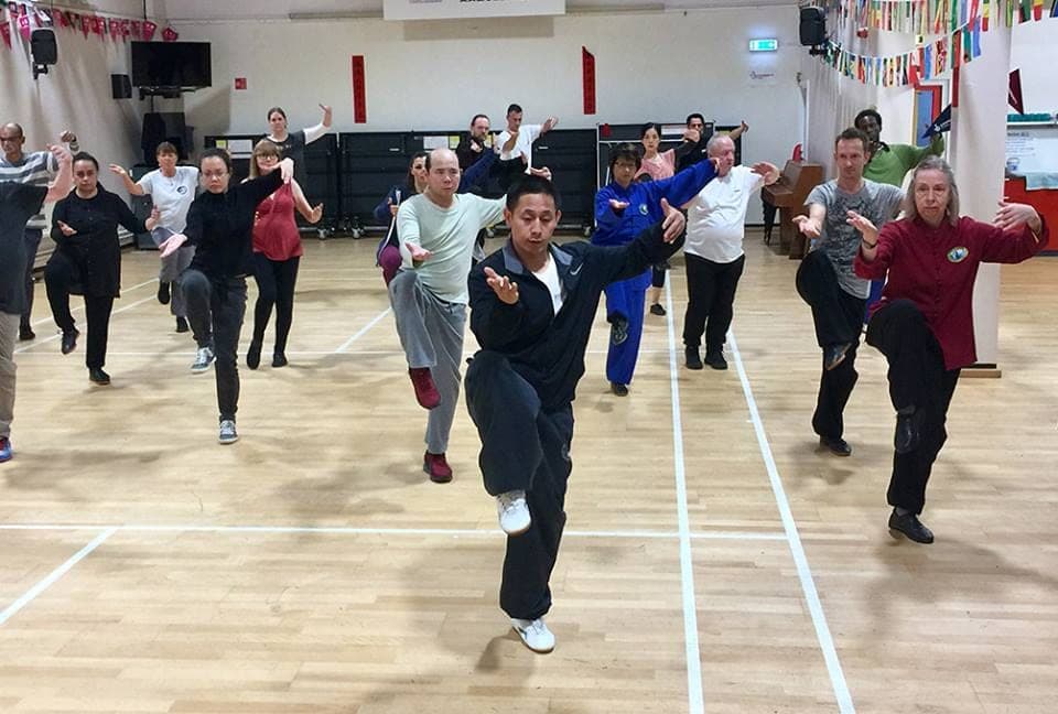 A class practises a form movement on one leg, knee raised, across a sports hall