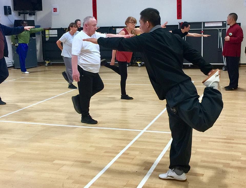 A teacher kicks one leg high to shoulder height during a warm-up while students balance behind him