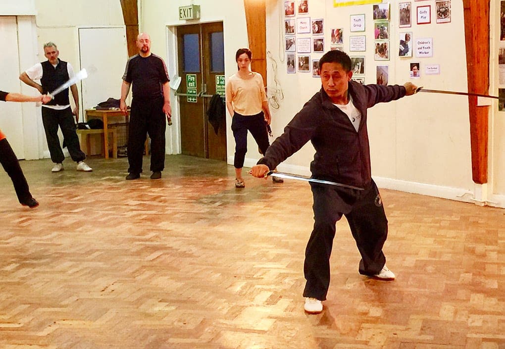 A teacher in a low stance holds two straight swords extended to either side in a church hall