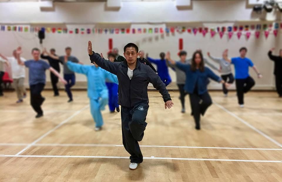 A teacher balances on one leg during a warm-up, with the class copying him across a sports hall